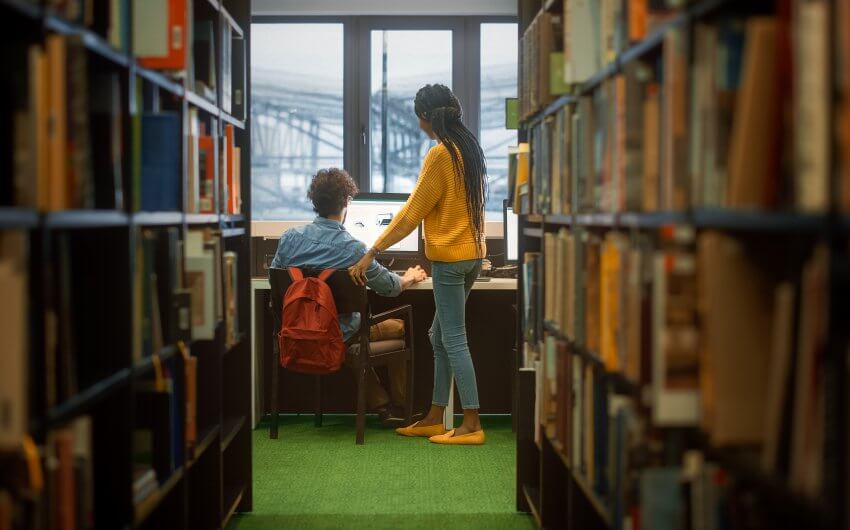 University Library: Boy Uses Personal Computer at His Desk, Talks with Girl Classmate who Explains, Helps Him with Class Assignment. Focused Students Study Together. Shot Between Rows of Bookshelves